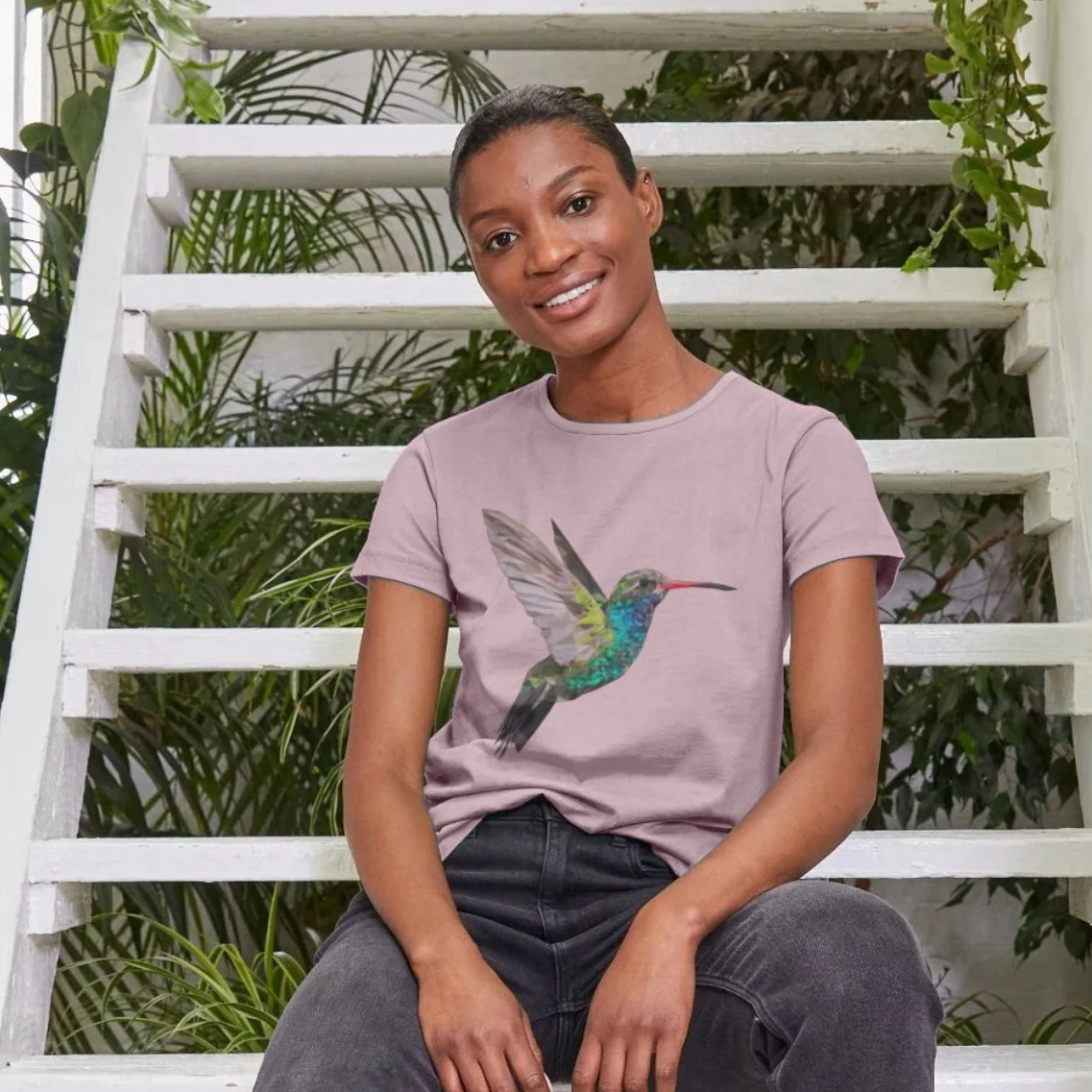 black woman sitting on ladder wearing a mauve t-shirt with a hummingbird motif by lumimango